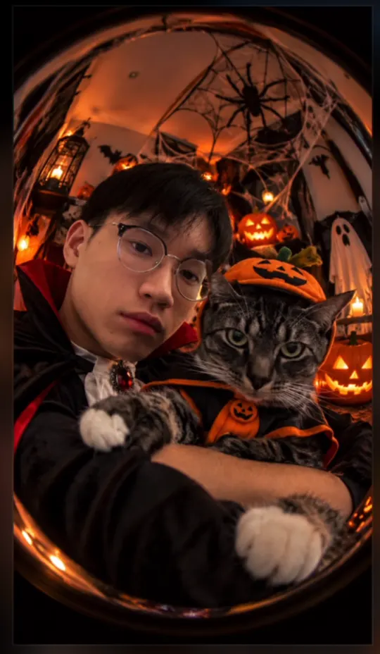 ChatGPT 輸入 /fisheye halloween 的結果：魚眼鏡頭下穿吸血鬼斗篷的人和戴南瓜帽的貓，背景是南瓜燈和蜘蛛網